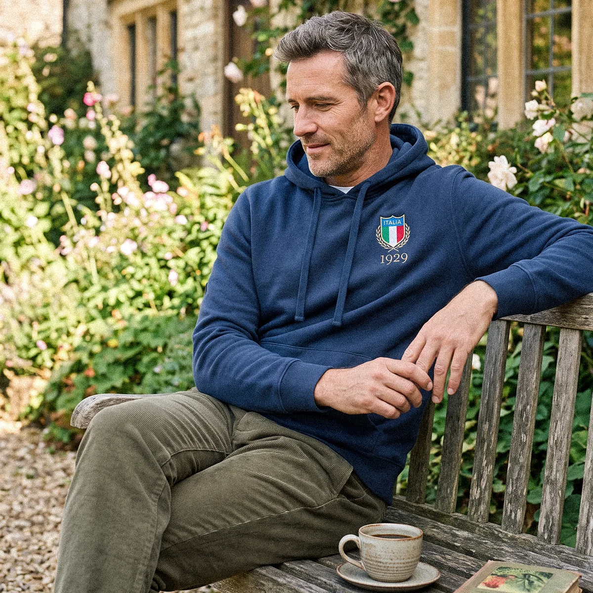 Rugby 1823 Pure Cotton Hoodie A man sits on a wooden bench outside a country cottage with a sunny garden behind him. He looks wistfully toward the ground.